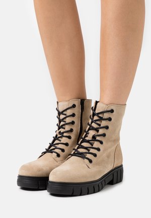 Botas de tobillo de ante beige con cordones negros y cremalleras laterales, con una suela texturizada y diseño de punta redondeada.
