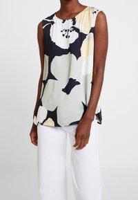Blusa senza maniche floreale in verde, nero e giallo. Tessuto morbido con scollo rotondo, vestibilità rilassata e orlo curvo, abbinata a pantaloni bianchi.