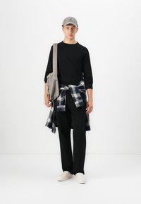 Un jeune homme portant une chemise noire à manches longues, un pantalon noir, une chemise à carreaux nouée autour de la taille, une casquette grise, des chaussures beiges et un sac à bandoulière, se tenant contre un fond blanc.