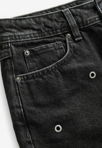 Gros plan sur un jean en denim noir montrant la poche avant, la boucle de ceinture, les rivets métalliques et les œillets argentés sur le tissu.