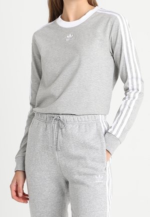 Kobieta ubrana w jasnoszary dres Adidas z białymi paskami na rękawach i spodniach, elastyczny pas z sznurkiem.