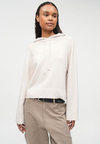 Gai rozā adīta kapuce ar ribinātu apakšmalu un piedurknēm, ar priekšējo kabatu un auklu kapuci, apvienota ar bēšiem biksēm un melnu jostu.