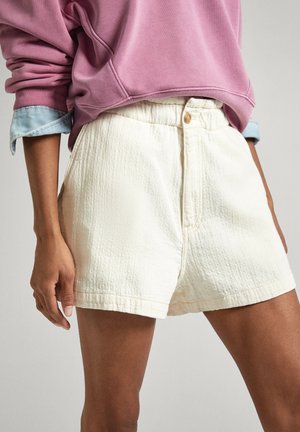 Person iført teksturerede off-white shorts med knap og lynlås, kombineret med en lilla sweatshirt og opkrogede ærmer på en denimskjorte.