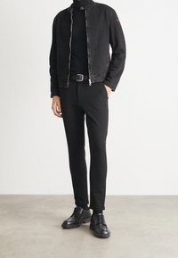 Czarna kurtka dżinsowa z wysokim kołnierzem i zamkiem błyskawicznym, w połączeniu z czarnym golfem, czarnymi spodniami o wąskim kroju i czarnymi butami.