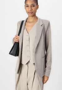 Beige Fischgrät-Blazer mit aufgesetzten Revers, kombiniert mit einer passenden Weste mit fünf Knöpfen und weit geschnittenen Hosen. Schwarze Handtasche als Accessoire.