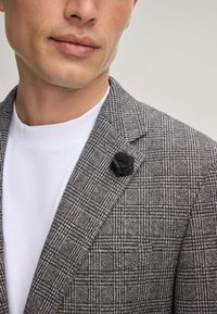 Grijze geblokte blazer met een notch lapel, gecombineerd met een witte t-shirt. Bevat een zwarte stoffen roos gespeld op de revers.