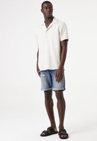 Lichtgekleurde linnen shirt met korte mouwen, blauwe versleten denim shorts en zwarte sandalen. Eenvoudig ontwerp, relaxte pasvorm en opgerolde shortszoom.