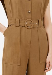 Brun jumpsuit med ett b älte vid midjan med en cirkulär spänne, knäppning framtill och stora sidofickor med en texturerad tygfinish.