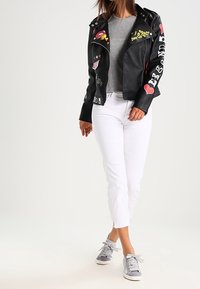 Kvinna i svart läderjacka med färgglada lappar, grå t-shirt, vita cropped byxor och gråa sneakers mot vit bakgrund.