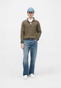 Pulôver de malha verde-azeitona com fecho de correr de um quarto e riscas horizontais. Boné azul, camisa riscada, jeans de corte flare azul claro e ténis brancos.