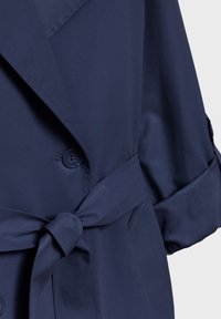 Marineblauwe trenchcoat met een ceintuur in de taille, grote revers en knoopsluiting. Gemaakt van een zachte, gestructureerde stof met opgerolde mouwen.