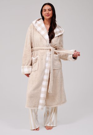 Femme portant un peignoir beige et duveteux avec une bordure à carreaux et une ceinture, debout pieds nus avec un pantalon de pyjama rayé sur un fond neutre.