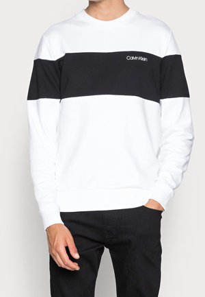 Mann trägt weißen Pullover mit schwarzem horizontalem Streifen und Calvin Klein Logo auf der Brust, kombiniert mit schwarzen Hosen.