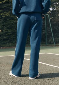 Türkisfarbene Sweatpants mit geradem Schnitt aus weichem Stoff, versehen mit weißen Seitenstreifen, kombiniert mit weißen Sportschuhen auf strukturiertem Boden.