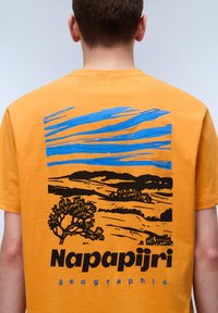 Oranssi puuvillainen t-paita, jossa sininen ja musta graafinen kuva kukkuloista ja puista. "Napapijri" -logo painettuna paksuilla mustilla kirjaimilla alapuolelle.