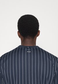 Mörkblå kortärmad skjorta med vertikala vita ränder, Fila-logotyp vid bakre halsringning, rund krage och strukturerad textil.