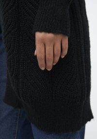 Sort strikket cardigan med et struktureret, mønstret design. Ærmekraven er ribbet, og en hånd griber om stoffet, hvilket viser dets tykkelse.