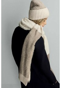 Bonnet en tricot beige et épais foulard, présentant des textures douces et moelleuses avec des motifs à blocs de couleurs en crème et beige, portés sur un vêtement noir.