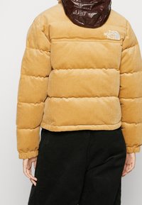 Person som bär en beige kordflors pufferjacka med en brun huva och svarta byxor, visande baksidan och sidan av jackan.