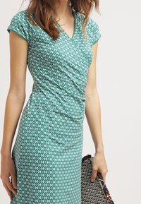 Nainen, jolla on päällään tiukka vihreä mekko, jossa on valkoisia ympyräkuvioita, pitää musta-valkoista kuviollista clutch-laukkua valkoista taustaa vasten.