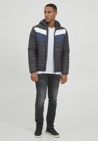 Veste rembourrée de base grise, avec des rayures sur la poitrine bleu marine et blanches. Comprend une capuche et une fermeture éclair à l'avant. Associée à un jean foncé et des baskets noires.
