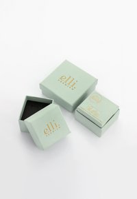 Cajas de cartón verde menta con texto dorado, una parcialmente abierta para revelar un interior negro. Diseño simple y limpio sin patrones o adornos adicionales.