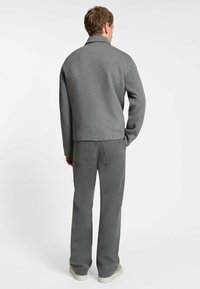 Mand vendt væk iført en grå langærmet jakke, grå bukser og hvide sneakers mod en ensfarvet hvid baggrund.