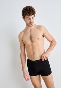 Black boxer shorts with a red "HUGO" logo on the elastic waistband, made from a smooth, stretchy material. Model has a neutral pose.