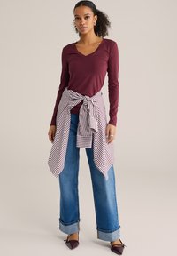 Bordová blúzka s dlhým rukávom a výstrihom do V v kombinácii s vysokými modrými džínami s manžetou na spodnom okraji a páskovou košeľou s pruhmi. Špicaté bordové balerínky.