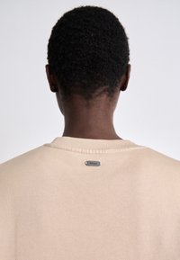 Beige sweatshirt with a crew neckline, featuring ribbed trim and a small silver logo tag on the back. Soft texture, casual design.