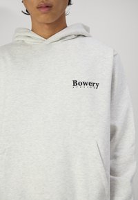 Grå hættetrøje lavet af blødt stof, med en kængurulomme og et logo i sort tekst på venstre bryst med teksten "Bowery Atelier."