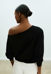 Maglione nero off-shoulder con maniche lunghe e ampie e orlo a costine. Indossato con pantaloni bianchi a vita alta. Texture liscia e morbida.