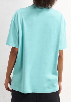 Personne aux cheveux bouclés portant un t-shirt ample à manches courtes bleu clair, vue de dos sur un mur blanc uni.