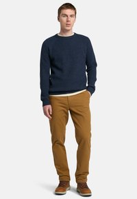 Maglione a coste blu navy con collo a giro, indossato sopra una camicia crema, abbinato a pantaloni marroni e stivali marroni con lacci.