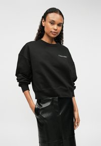 Μαύρη βαμβακερή μπλούζα με χαμηλούς ώμους και ριμπ μανσέτες. Διαθέτει λευκό λογότυπο "Calvin Klein" στο στήθος. Συνδυάζεται με μια γυαλιστερή μαύρη φούστα.