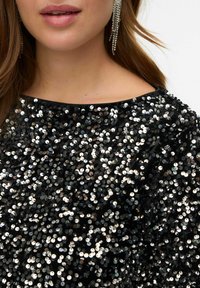 Crni materijal prekriven reflektirajućim šarenim šljokicama, stvarajući sjajnu teksturu. Ima okrugli izrez i uparen je sa srebrnim visećim naušnicama.