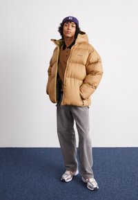 Tan puffjakke med hætte, lynlås foran og grøn foring. Bæres over en brun polo og grå fløjlbukser. Sølv sneakers fuldender looket.