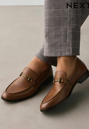 Barna bőr mokaszinok aranyszínű fém díszítéssel, szürke kockás, testreszabott nadrággal világos padlón.