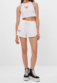 Bílý žebrovaný crop top a odpovídající šortky s červeným textem "MALIBU". Obsahuje stahovací pas a volný střih. Doplněno o černé platformové boty.