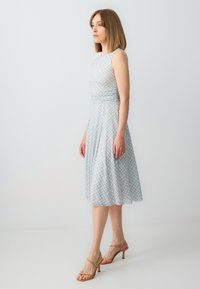 Helles blaues, ärmelloses Kleid mit tailliertem Oberteil und ausgestellt Rock, der mit einem karierten Muster in gelben und dunkleren Blautönen versehen ist. Sandalen mit Absatz.