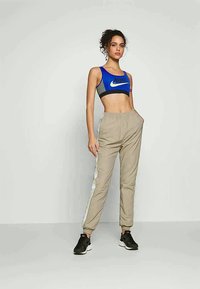 Bustier sportiv albastru și gri cu un logo, pantaloni jogger bej cu panouri laterale albe și pantofi sport negri cu un talpă texturată.