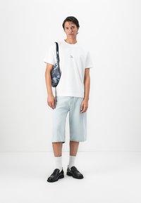 Homem a usar uma t-shirt branca, shorts de ganga claros, sapatos loafers pretos, meias brancas, e a carregar uma mala de ombro com padrão azul em fundo branco.