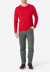 Rotes Langarm-Henley-Shirt, weiche Textur, kombiniert mit grünen Cargo-Hosen mit Seitentaschen und braunen Akzenten. Schwarze Schnürstiefel.