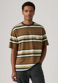 Camiseta marrón a rayas con mangas cortas, que presenta líneas horizontales en color crema y verde oscuro. Incluye un pequeño bolsillo en el lado izquierdo del pecho con el logo.