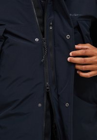 Marinblå isolerad jacka med dragkedja fram, tryckknappar och texturerat tyg. Har kontrasterande sömmar och en närbild av handjustering.