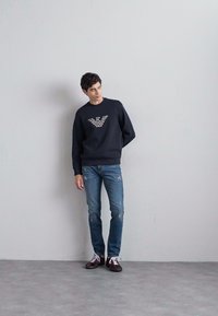 Zwarte sweatshirt met een gepatternde logo op de borst, gecombineerd met vervaagde blauwe jeans. Bruine schoenen zichtbaar, staande tegen een lichte muur.