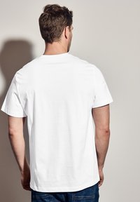 Valkoinen puuvillapaita, jossa on pyöreä pääntie ja lyhyet hihat. Paidan takana ylemmässä osassa on hienovarainen logo vaaleassa painatuksessa.