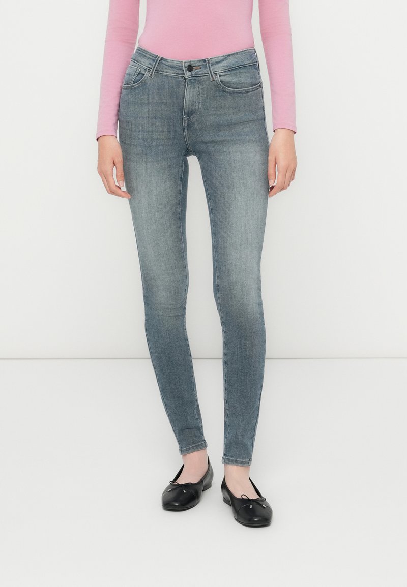 Egy nő fakó szürke skinny farmernadrágot, hosszúujjú rózsaszín felsőt és fekete balettcipőt visel, háttérben egy fehér fal.