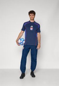 Marine sportshirt met rode accenten en logo, gecombineerd met bijpassende marine joggingsbroek. Speler houdt een blauw-witte voetbal vast.
