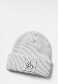 White ribbed knit beanie with folded cuff and patch reading “Lächle! Du kannst sie nicht alle töten” with a smiley face above text.